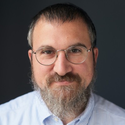 zensmart is showing A man with glasses, a short dark beard with some gray, and short dark hair smiles softly. He is wearing a light blue, collared shirt and is posed against a plain, dark background. with print workflow automation