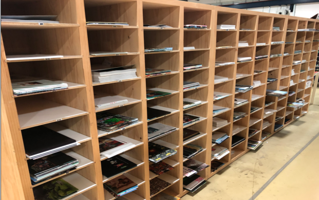 zensmart is showing Wooden shelving units with multiple cubbyholes, each containing stacks of magazines, papers, and booklets organized by slot, standing on a light-colored floor in an indoor setting. with print workflow automation