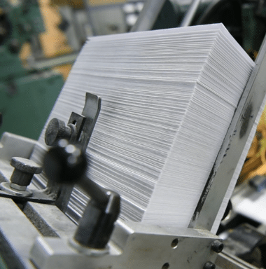 zensmart is showing A close-up view of a stack of white paper sheets loaded in a printing press, ready for processing. The machinery's metal parts and adjustment knobs are visible in the foreground. with print workflow automation