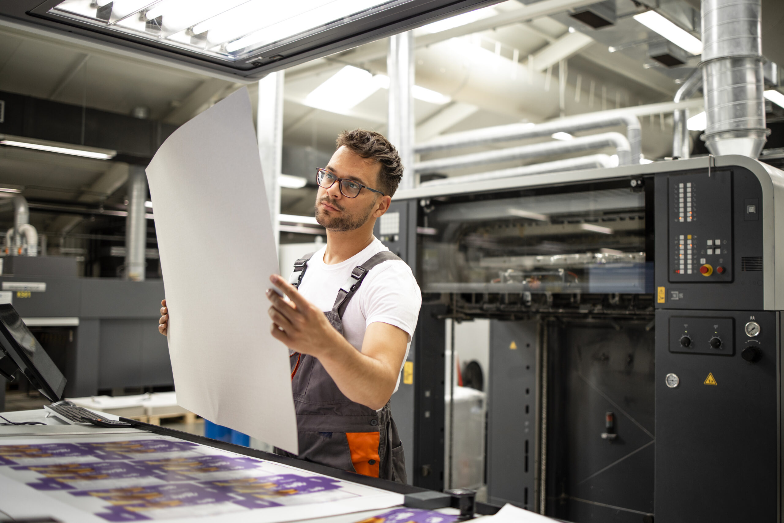 zensmart is showing A man in overalls and glasses inspects a large printed sheet in a modern printing facility, standing next to industrial printing equipment with control panels and printed materials visible. with print workflow automation