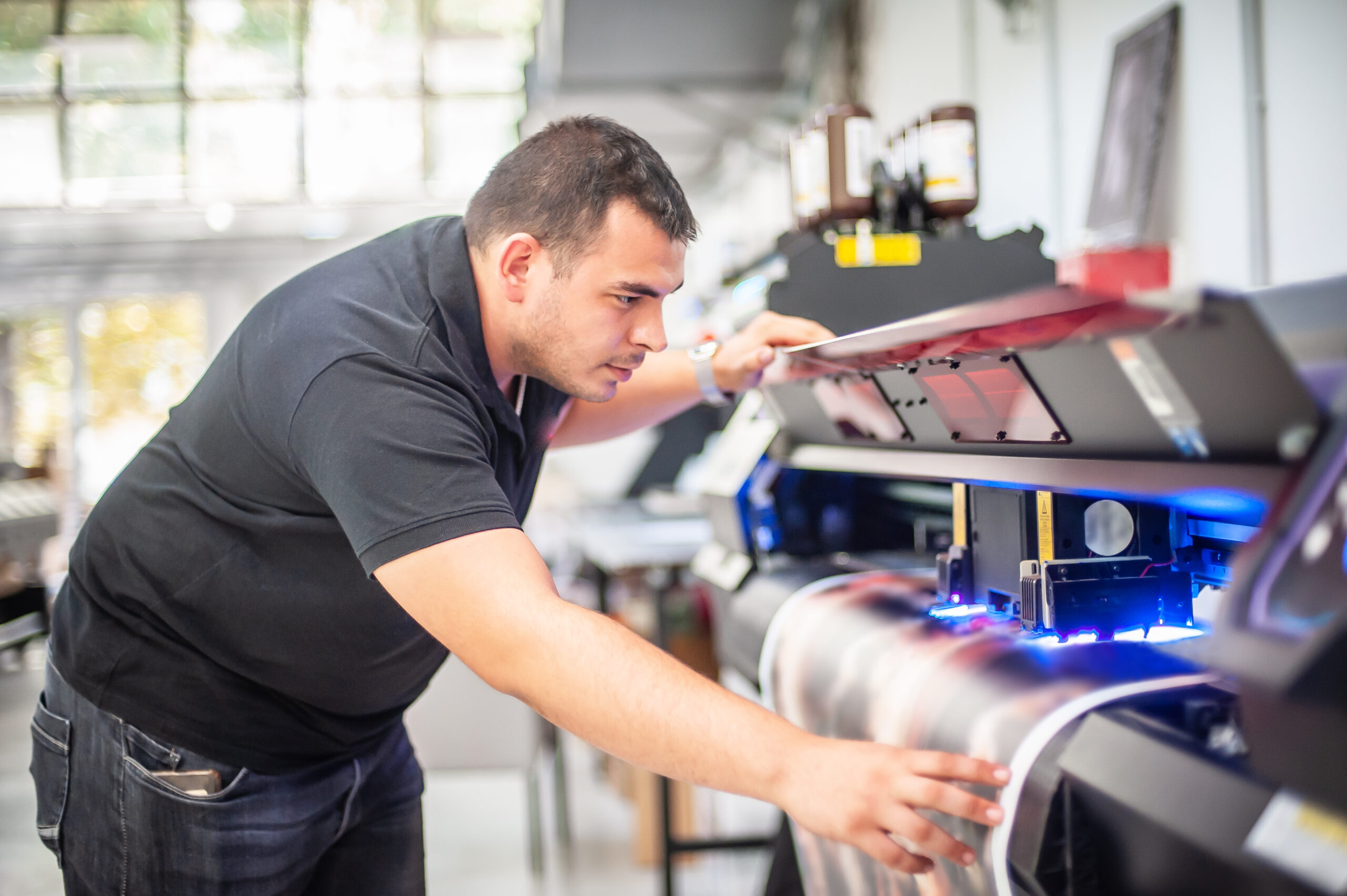 zensmart is showing A man in a black polo shirt inspects a large-format printer as it prints a colorful image on paper in a bright, modern workspace. with print workflow automation