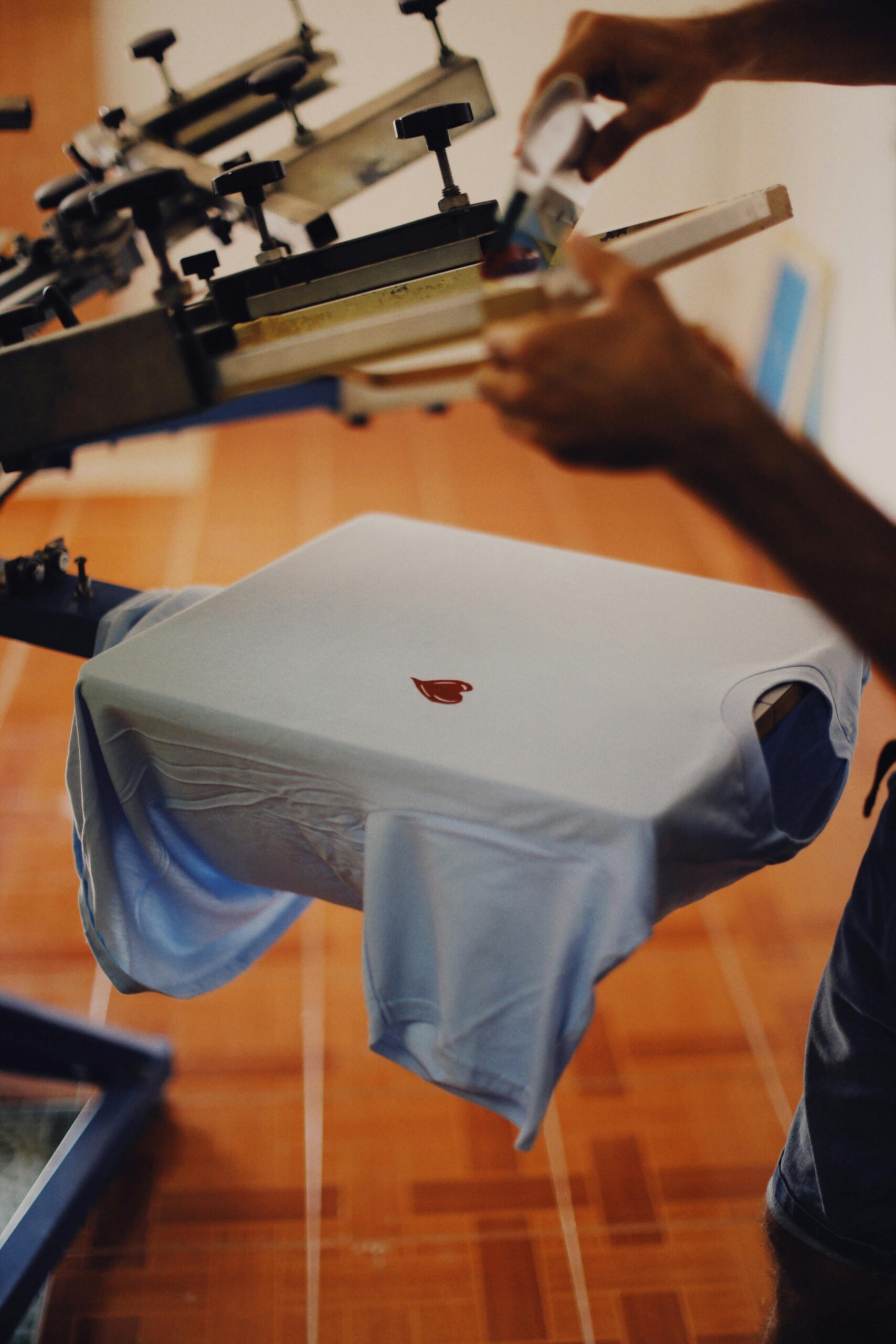 zensmart is showing A person at a Screen Shop uses a screen printing machine to print a small red heart design onto a light blue t-shirt, which is stretched out on the machine. with print workflow automation